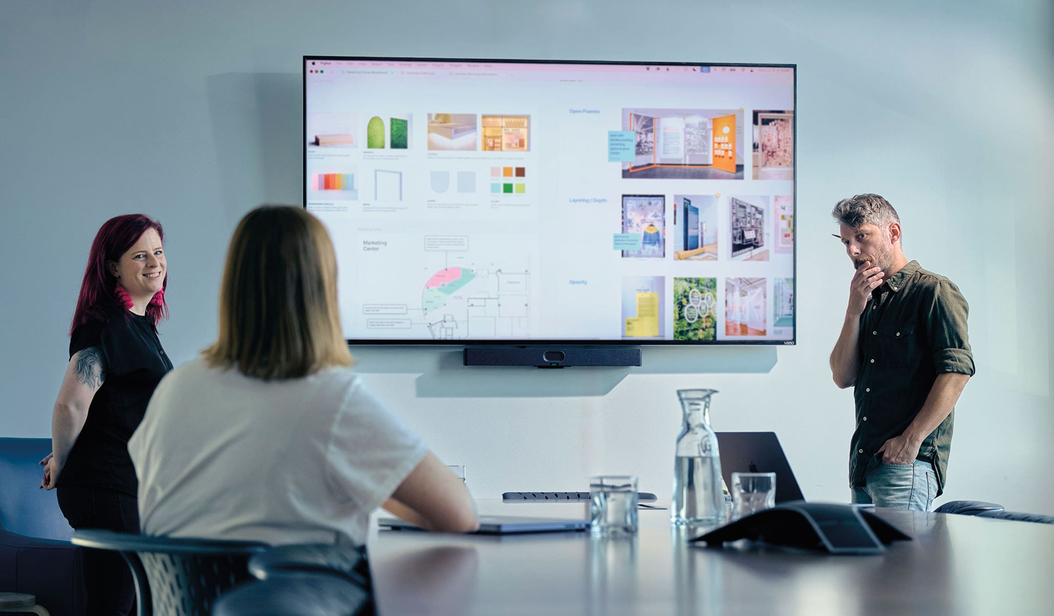 A white man with a beard and a white woman with red hair present a digital mood board, listening to a woman with her back to the camera.